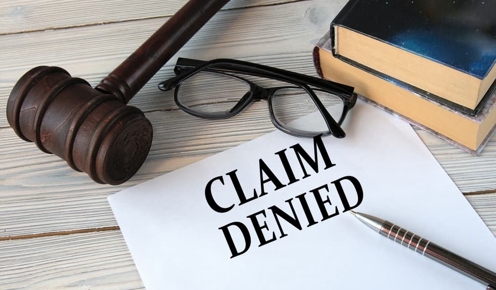 The words "CLAIM DENIED" printed on white paper, set against a background featuring a judge's gavel, glasses, and a pen.